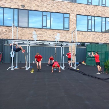 A group of people in red shirts engage in various fitness exercises outdoors, using equipment such as pull-up bars, battle ropes, and resistance bands, with a brick building in the background.