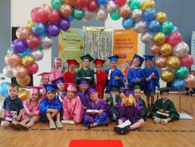 A group of children in graduation gown are under a arch of rainbow balloons at the YMCA Nursery