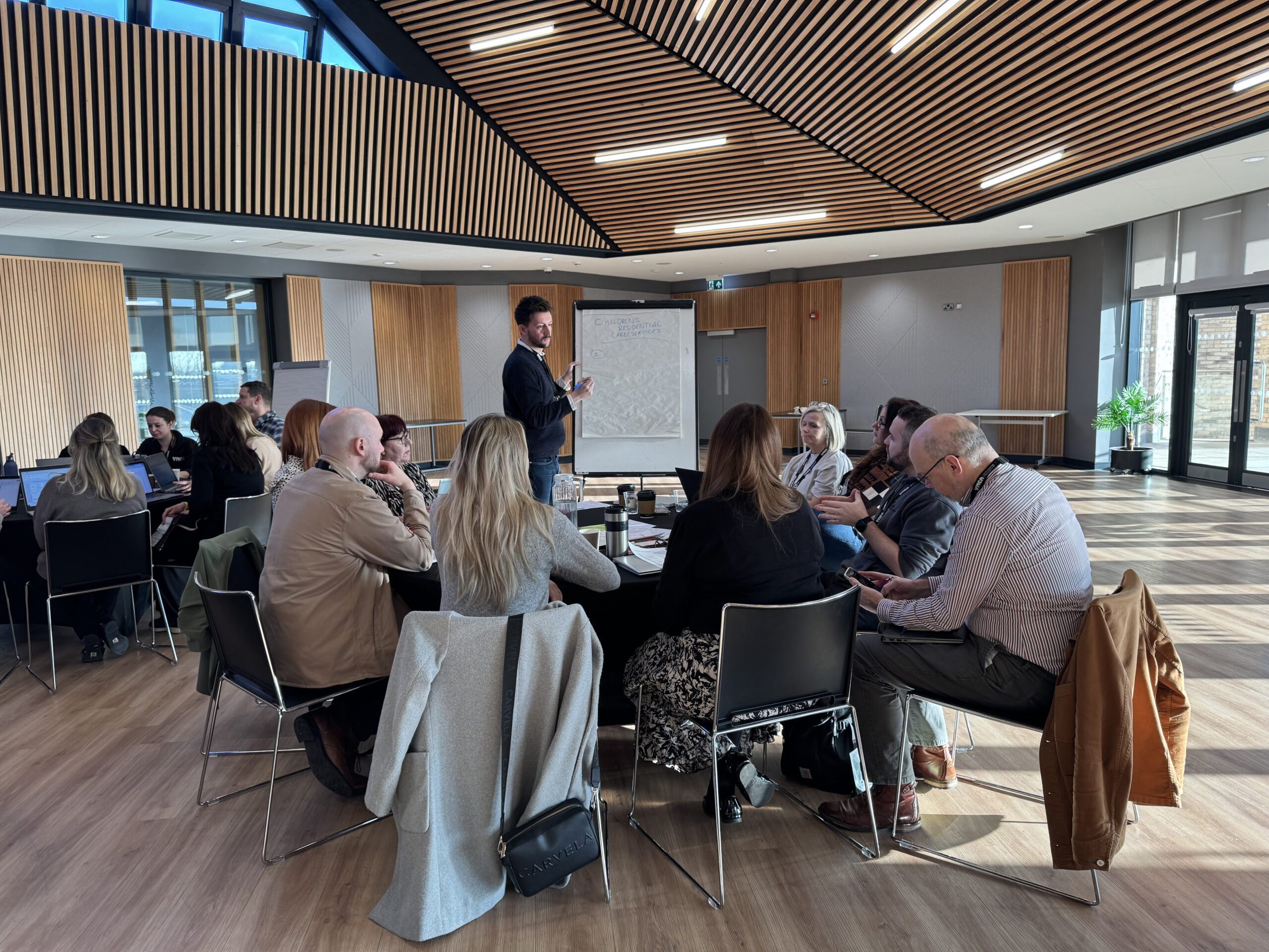 This photo shows a roundtable discussion at the YMCA Village Conference Centre in Newark. The modern venue features wooden slat ceilings, skylights, and a professional setup with attendees seated in groups, a presenter, and a large screen.