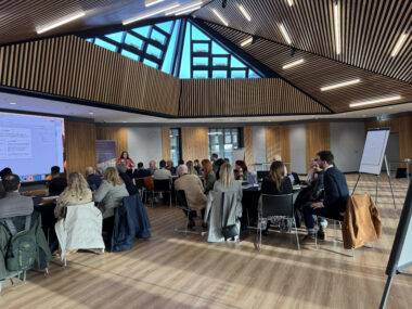This photo shows a roundtable discussion at the YMCA Village Conference Centre in Newark. The modern venue features wooden slat ceilings, skylights, and a professional setup with attendees seated in groups, a presenter, and a large screen.