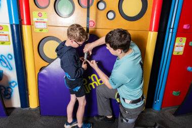 YMCA Y-kids - instructor coaching young boy how to attach a climbing harness in Clip n' Climb Centre, Newark.