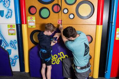 YMCA Y-kids - instructor coaching young boy how to attach a climbing harness in Clip n' Climb Centre, Newark.